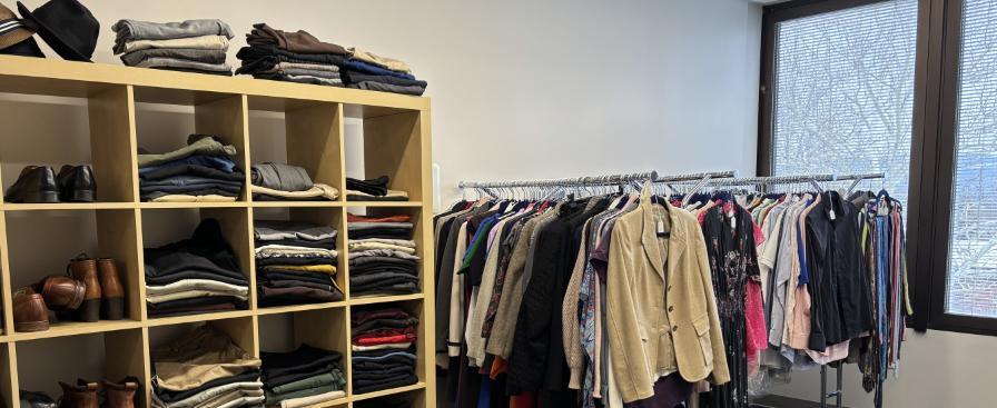 A small room containing neatly organized professional clothing. On the left, an open wooden shelving unit holds folded pants, sweaters, and several pairs of shoes. To the right, three clothing racks display a variety of jackets, shirts, and other garments on hangers. Large windows in the background let in natural light, and part of a table is visible in the foreground.
